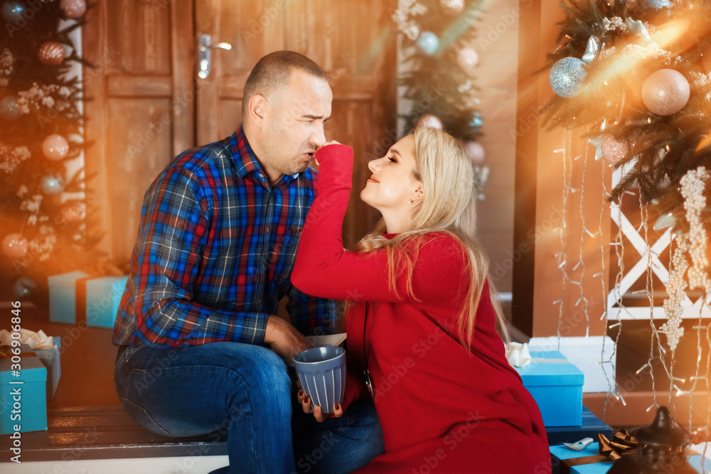 Couple sits on christmas decorated porch with grey cups of tea, coffee, hot wine in cup. Blonde woman in red dress smiles and touches man's nose, man in plaid shirt wrinkles face.