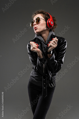 Slim caucasian girl with rough facial features in a black jacket listen to music with big earphones isolated on black background