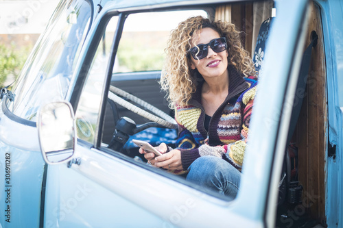 Cheerful people and happy beautiful curly blonde young caucasian woman type a message at the phone sit down inside an old vintage cute van -concept of travel driver and trendy fashion style