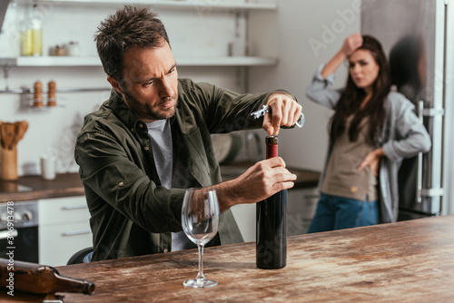 Selective focus of alcohol depended man opening wine bottle and upset wife on kitchen