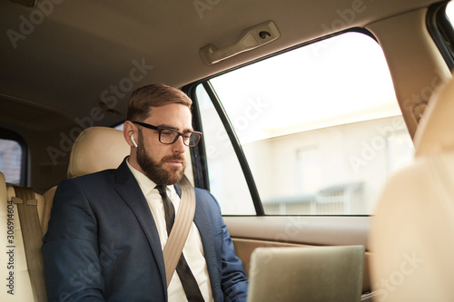 Young businessman in eyeglasses sitting on back seat of the car and working on laptop computer © AnnaStills