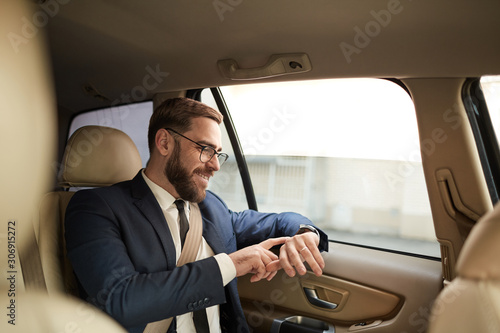 Happy young businessman in eyeglasses sitting on back seat and looking at his wristwatch while travelling by taxi © AnnaStills