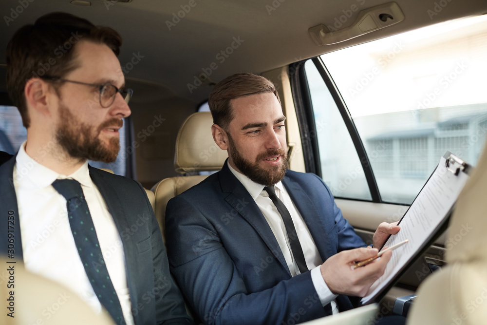 Young businessman pointing at business contract and discussing it with his colleague while they sitting on back seat of the car