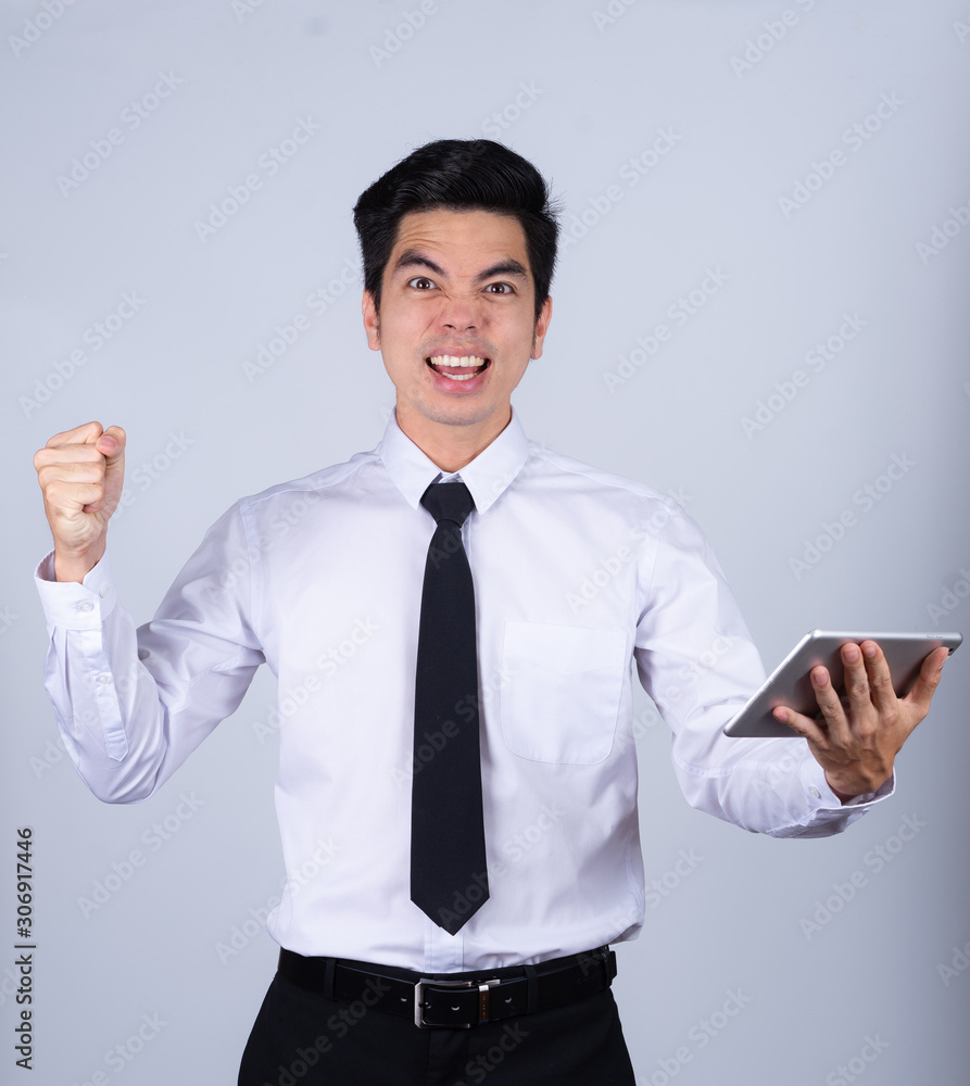 Portrait handsome young asian man wearing a white shirt holding smart phone or tablet excitement or celebrating his victory sign isolated on gray background in studio. Asian man people. Portrait handsome young asian man wearing a white shirt holding smart phone or tablet excitement or celebrating his victory sign isolated on gray background in studio. Asian man people.