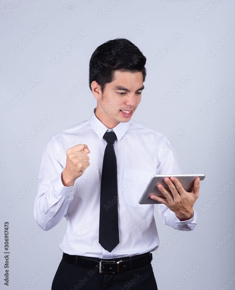 Portrait handsome young asian man wearing a white shirt holding smart phone or tablet excitement or celebrating his victory sign isolated on gray background in studio. Asian man people. Portrait handsome young asian man wearing a white shirt holding smart phone or tablet excitement or celebrating his victory sign isolated on gray background in studio. Asian man people.