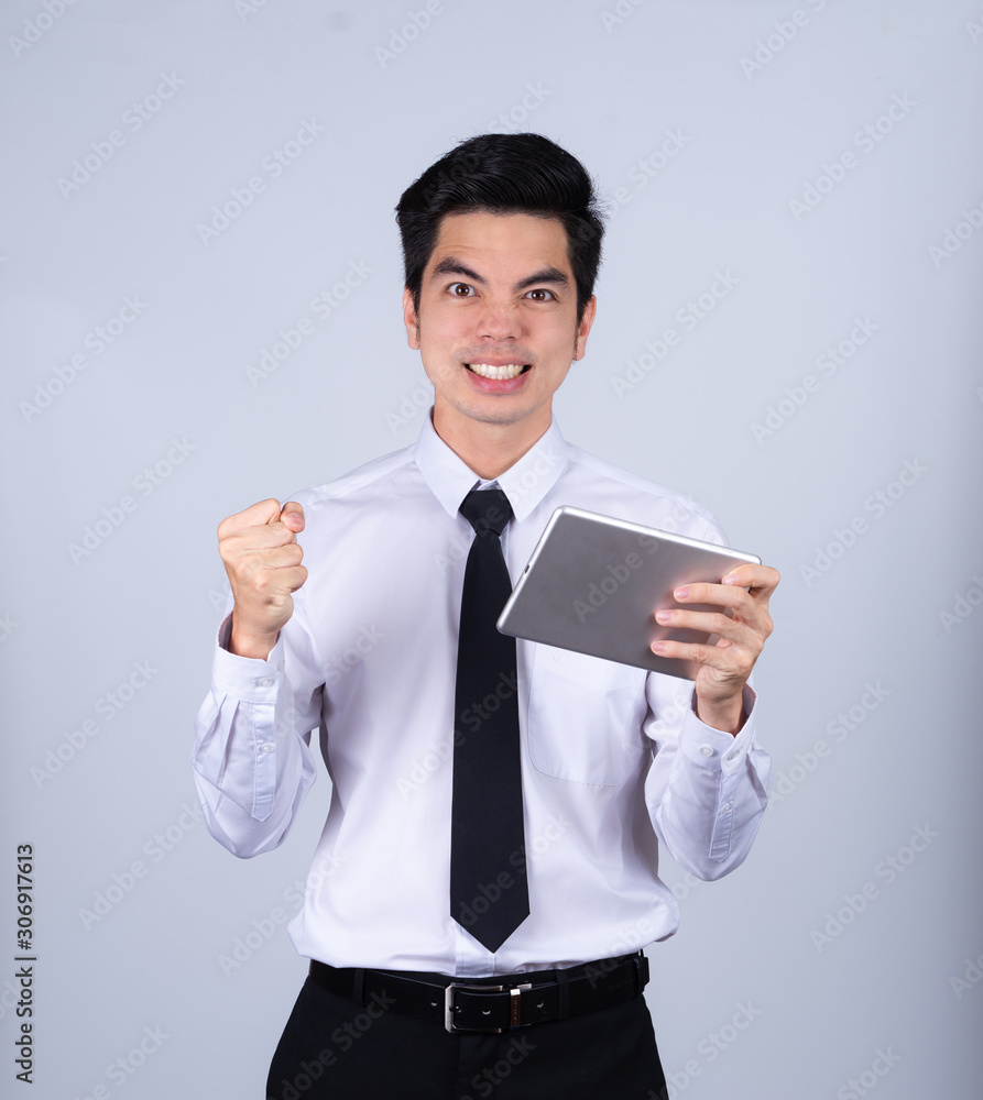 Portrait handsome young asian man wearing a white shirt holding smart phone or tablet excitement or celebrating his victory sign isolated on gray background in studio. Asian man people. Portrait handsome young asian man wearing a white shirt holding smart phone or tablet excitement or celebrating his victory sign isolated on gray background in studio. Asian man people.