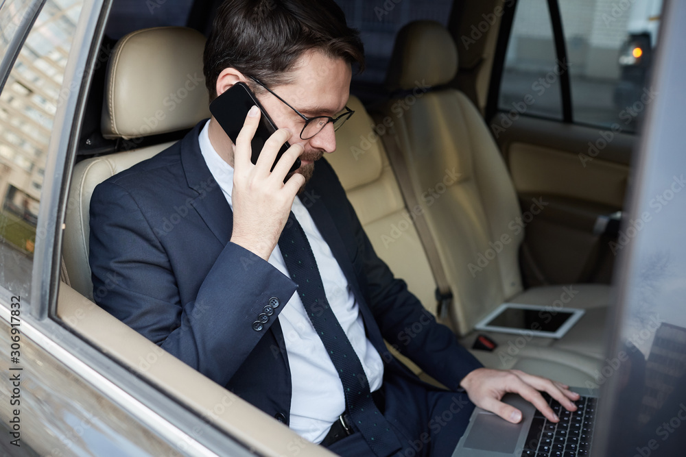 Young successful businessman in suit sitting on back seat of the car talking on mobile phone and typing on laptop during driving