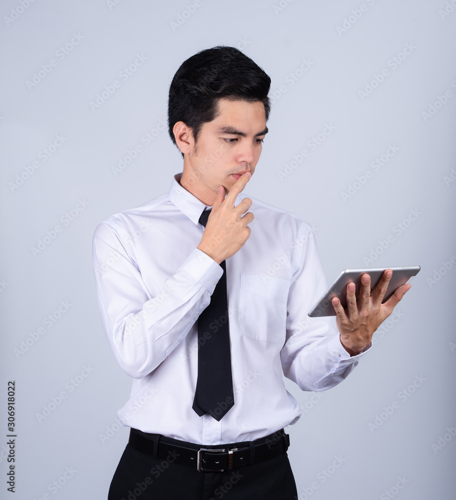 Portrait handsome young asian man wearing a white shirt holding smart phone or tablet thinking isolated on gray background in studio. Asian man people. business success concept. Portrait handsome young asian man wearing a white shirt holding smart phone or tablet thinking isolated on gray background in studio. Asian man people. business success concept.
