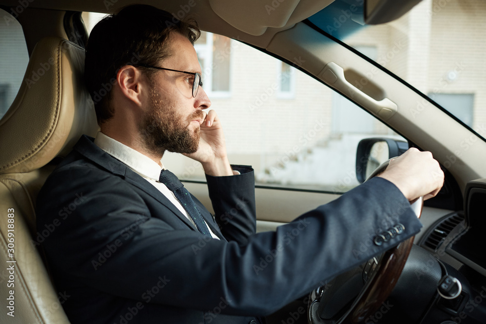 Side view of elegant driver in suit talking on mobile phone and watching for the road while driving the car