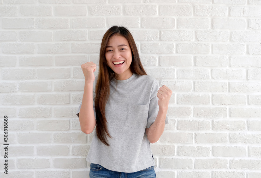 Portrait of young asian woman exciting and winning, Happy sucessful asia girl hand rised smiling and looking at camera while standing in fornt of white wall, People gesture in positive emotion Portrait of young asian woman exciting and winning, Happy sucessful asia girl hand rised smiling and looking at camera while standing in fornt of white wall, People gesture in positive emotion