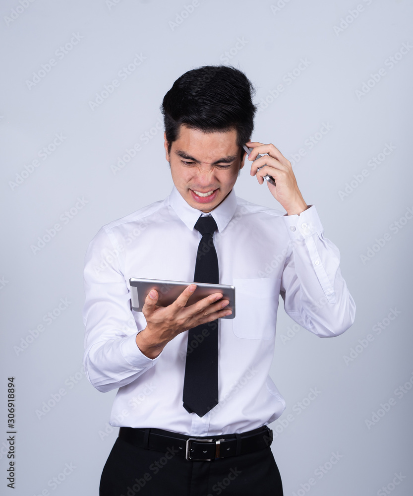 Portrait handsome young asian man wearing a white shirt holding smart phone or tablet stressed and anxiety isolated on gray background in studio. Asian man people. business success concept. Portrait handsome young asian man wearing a white shirt holding smart phone or tablet stressed and anxiety isolated on gray background in studio. Asian man people. business success concept.