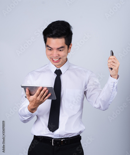 Portrait handsome young asian man wearing a white shirt holding smart phone or tablet excitement or celebrating his victory sign isolated on gray background in studio. Asian man people.