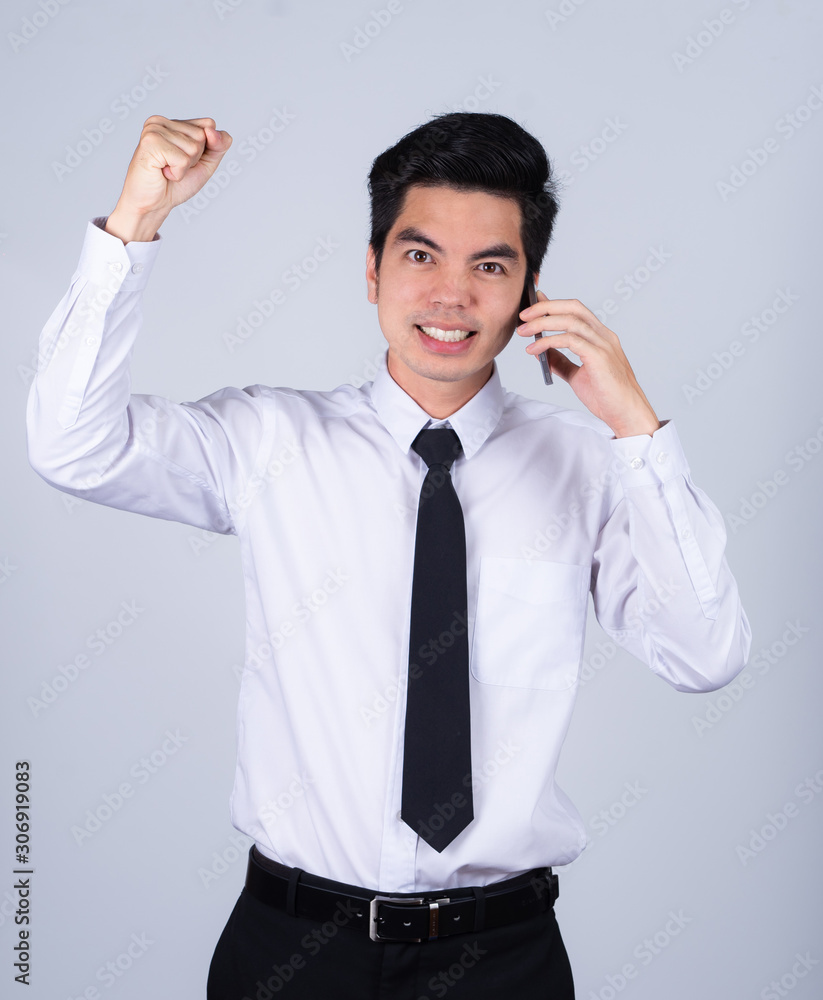 Portrait handsome young asian man wearing a white shirt holding smart phone excitement or celebrating his victory sign isolated on gray background in studio. Asian man people. Portrait handsome young asian man wearing a white shirt holding smart phone excitement or celebrating his victory sign isolated on gray background in studio. Asian man people.