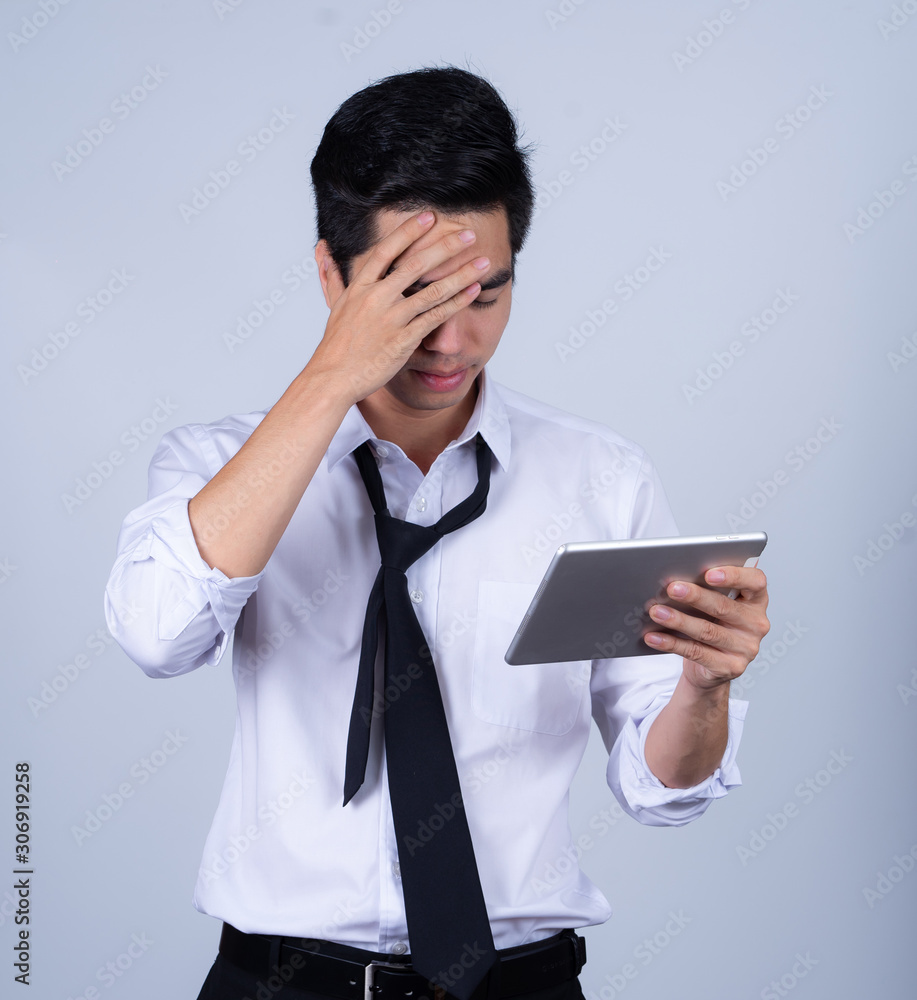 Portrait handsome young asian man wearing a white shirt holding smart phone or tablet stressed and anxiety isolated on gray background in studio. Asian man people. business success concept. Portrait handsome young asian man wearing a white shirt holding smart phone or tablet stressed and anxiety isolated on gray background in studio. Asian man people. business success concept.