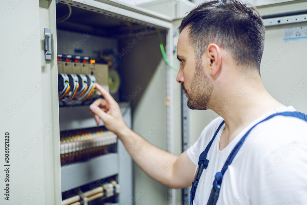 Focused handsome caucasian worker in overalls standing in front of transformer and checking on electricity.