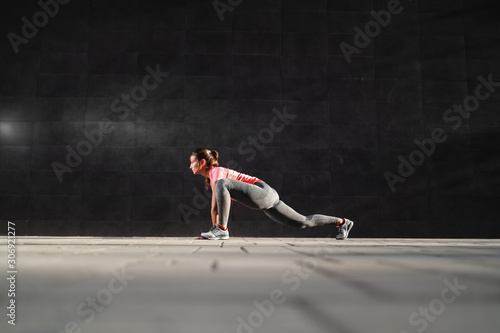 Side view of fit attractive caucasian brunette in sportswear and with ponytail stretching leg. In background is dark wall. © dusanpetkovic1