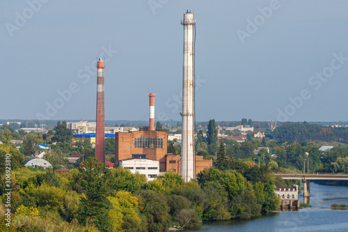 Red bricked thermal power station with tall exhaust pipes in trees with river, bridge and city at background on sunny day with blue sky. Industrial buildings and urban views concept