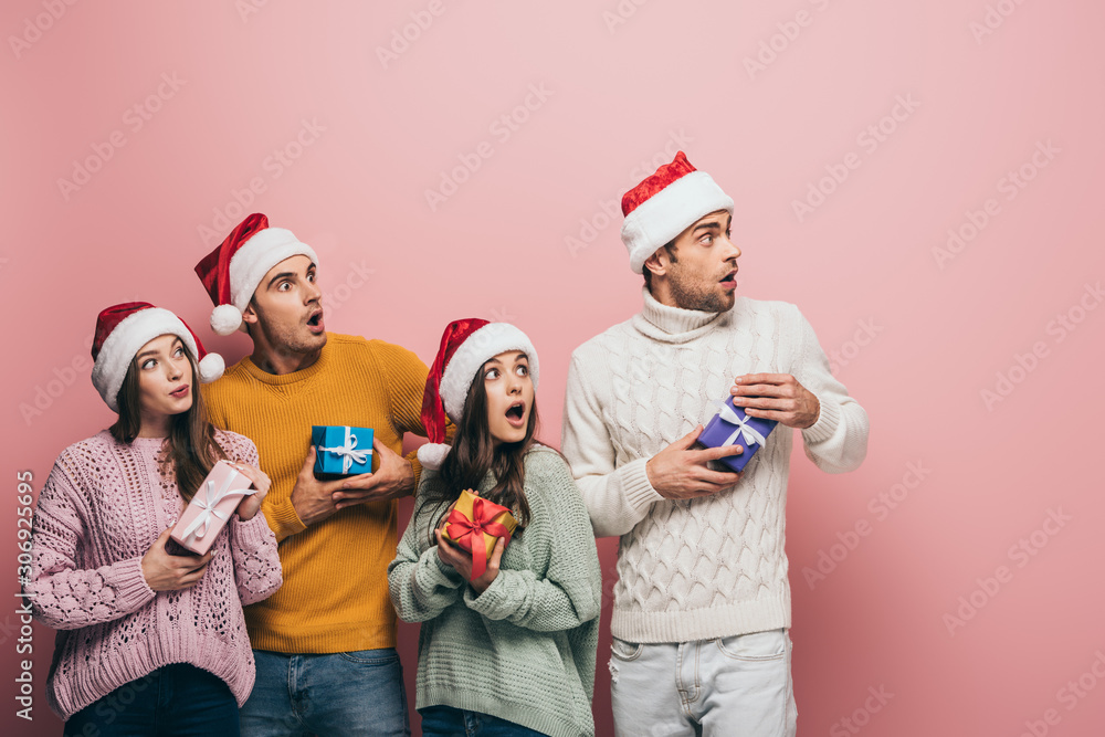Fototapeta premium shocked friends in santa hats holding christmas presents, isolated on pink