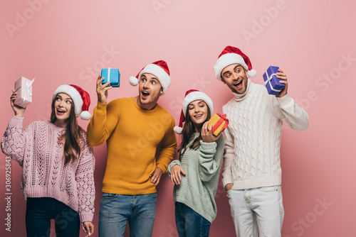 excited friends in santa hats holding christmas presents, isolated on pink