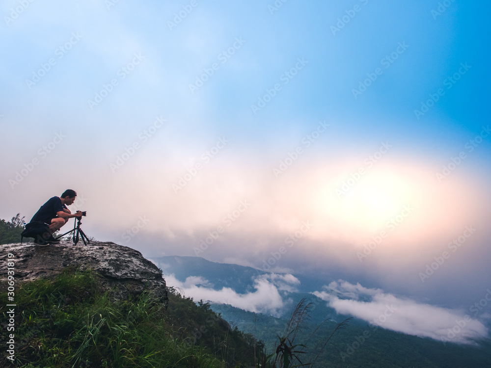 Nature photographer takes photos with camera on tripod at cliff. Dreamy foggy landscape. Morning sunrise  blue misty in beautiful valley below.