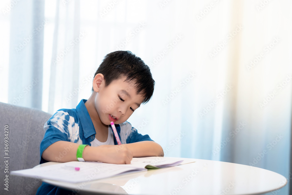 Asia boy wear white T-shirt and cover with two tone of shirt is doing homework. He used pencil and writing on a book. The boy is thinking, happily and funny, family and learning concept Asia boy wear white T-shirt and cover with two tone of shirt is doing homework. He used pencil and writing on a book. The boy is thinking, happily and funny, family and learning concept