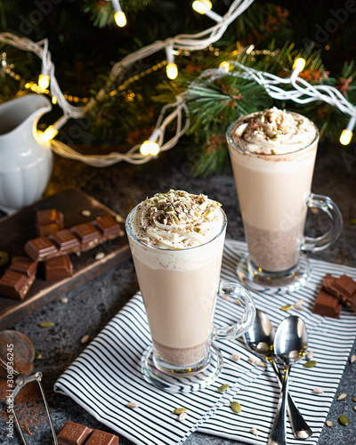 Christmas latte with whipped cream and pieces of chocolate with spoons on a napkin on a Christmas tree lighting background