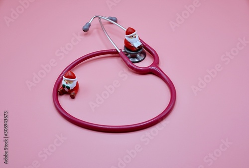 Two santa claus toys standing in front of a pink stethoscope on the pink background
