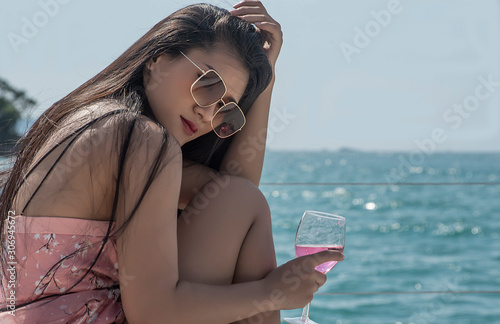 Beautiful Asian women sit and hold wide glasses, wear sunglasses, sunbathe on yachts during holidays.