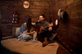 Young couple in love on romantic date in wooden room hut on christmas new years eve. Brunette man, wearing grey winter sweater, holding playing guitar for pretty woman covered with warm blanket.