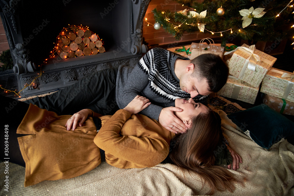 Handsome brunette man in grey sweater and young woman in mustard yellow knitted dress lying on the floor carpet near Christmas tree, embracing. Couple in love by fireplace in dark room with brick wall