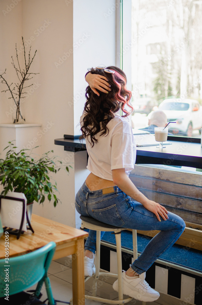 Stylish girl in a white T-shirt and jeans sits in a cafe on the bar stool. Beautiful ass of a young, white girl in tight blue jeans. Bright closeup photo.