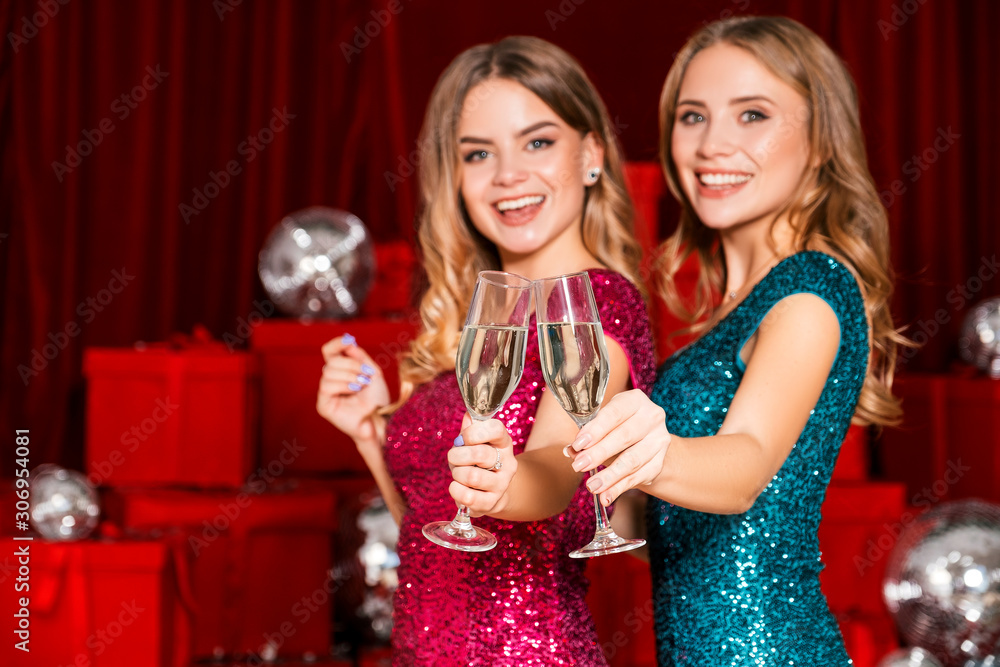Two cheerful happy girls having a new year party, celebrating christmas. They drink champagne and open presents. They wear sexy dresses that curves their body in sexy way. Many red presents on back Two cheerful happy girls having a new year party, celebrating christmas. They drink champagne and open presents. They wear sexy dresses that curves their body in sexy way. Many red presents on back