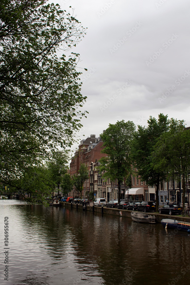 View of canal, parked boats and cars, trees and historical and traditional buildings in Amsterdam. It is a summer day with cloudy sky.