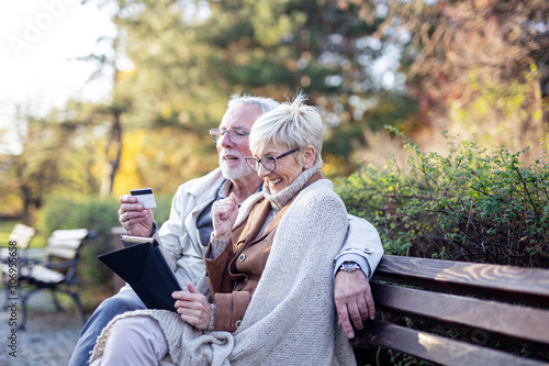 Senior couple sitting in the park on bench and shopping on line with tablet and credit card