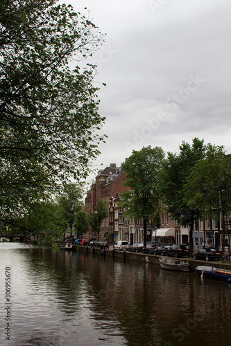 View of canal, parked boats and cars, trees and historical and traditional buildings in Amsterdam. It is a summer day with cloudy sky.