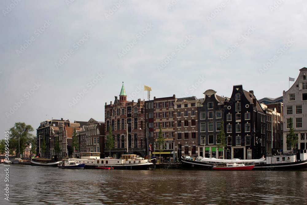 View of Amstel river, canal boats and historical, traditional and typical buildings reflecting Dutch architectural style in Amsterdam. It is a summer day with cloudy sky.