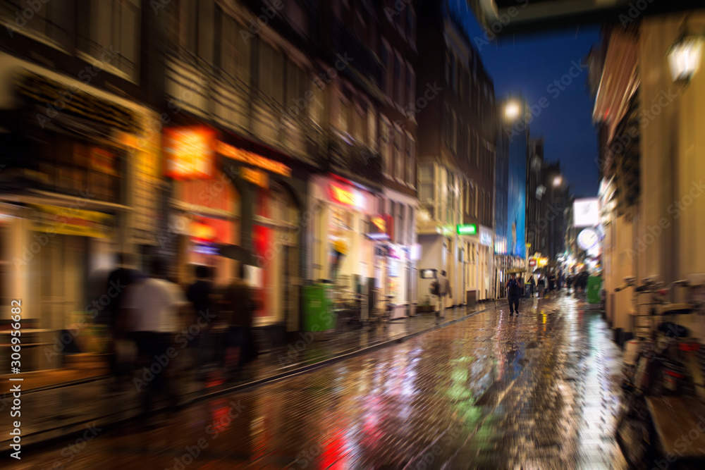 Blurry motion image of people walking on Warmoesstraat street in Amsterdam. It is one of the main shopping streets with cafes, restaurants and shops. It is a rainy summer night. Youth culture concept
