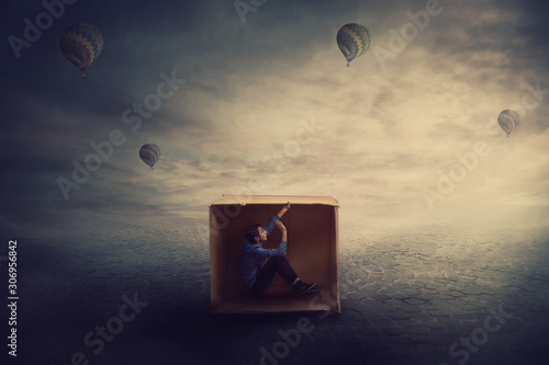 Surreal scene as a trapped guy want to get out of a cardboard package, needs to think outside the box. Scared man caged by own fears sitting inside a carton hut. Introvert and extrovert concept.