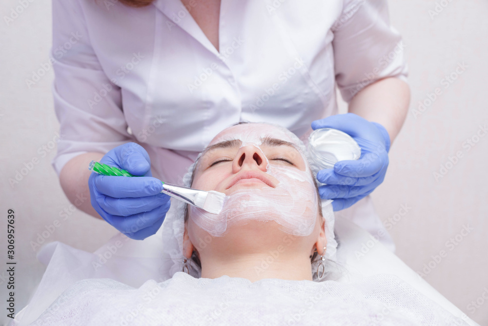 Crop master applying white mask with brush on face and neck of young woman in towel lying on table Crop master applying white mask with brush on face and neck of young woman in towel lying on table