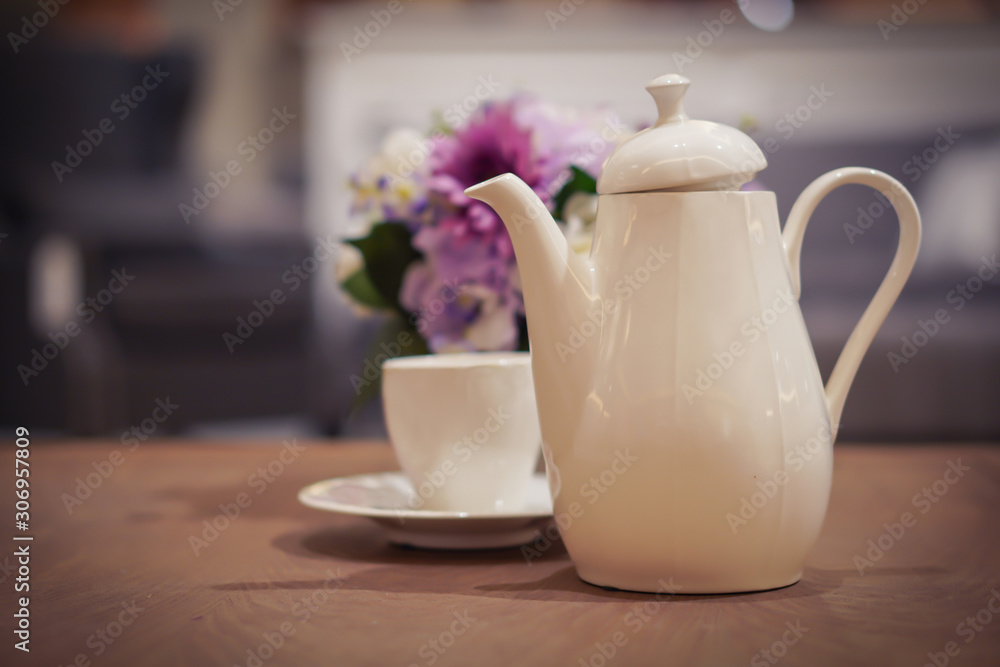 Focus selection: a white coffee pot placed on the table with a coffee cup and flowers as a blurred background Focus selection: a white coffee pot placed on the table with a coffee cup and flowers as a blurred background