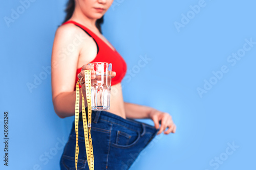 slim woman in big fat jeans show her losing weight and holdind glass of water with measure tape, selected focus, blue background.
