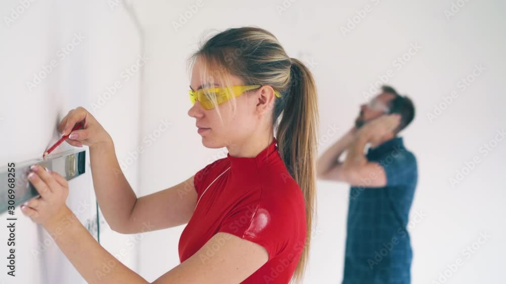 Dolly cam: pretty girl in yellow goggles checks wall level with modern instrument by young man listening to music in room Dolly cam: pretty girl in yellow goggles checks wall level with modern instrument by young man listening to music in room