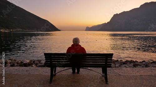 Canvas Print Lonely old man sits on a wooden bench at dusk looking at the lake and the light on the horizon