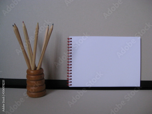 lápices de madera con minas de varios colores rojo, azul, verde, amarillo en porta lápices de madera marrón miel con ribetes circulares, con fondo gris, negro, y cuaderno de anillas rojas para anotar photo