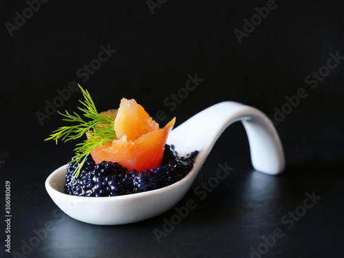 Black caviar with smoked salmon bite in white porcelain spoon over black background.
