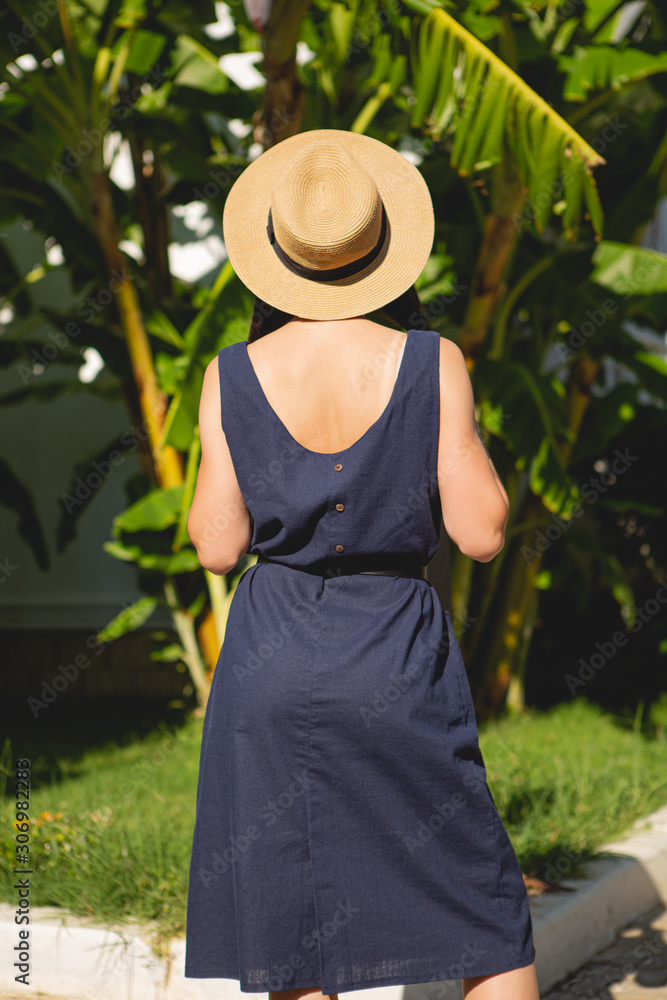 stylish young attractive woman dressed in wearing a blue short casual dress and hat photographed in the summer on a background of palm trees and the sea in the bright sun