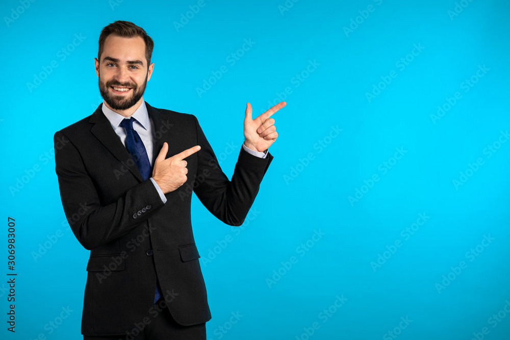 Happy smiling businessman in corporate suit presenting, showing something isolated on blue background. Portrait of man, he pointing with arms on his left with copy space.