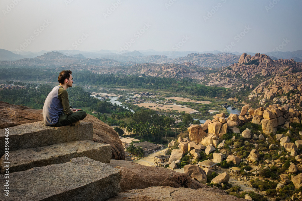 Tourist guy sitting on the edge of a hill. Beautiful view of the amazing Hampi's ruins from Matanga hill. Karnataka, India