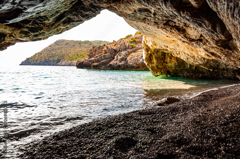 Amazing seascape view from unique sea cave overhanging above water on famous Cala Bianca beach scenic surroundings. Crystal clear sea water waves washing pebbles inside the massive rocky cave