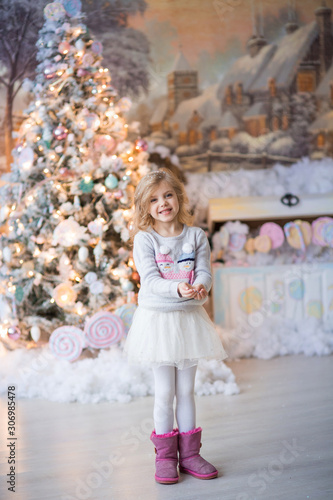 pretty happy blonde girl with curls of 6-7 years in a thematic holiday Christmas sweater with snowmen, holding snow in her hands, Christmas, near the New Year tree, winter © Евгения Янцева